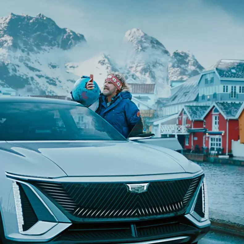 Will Ferrell standing outside of an electric car in a Scandinavian village with his hand punched through a globe