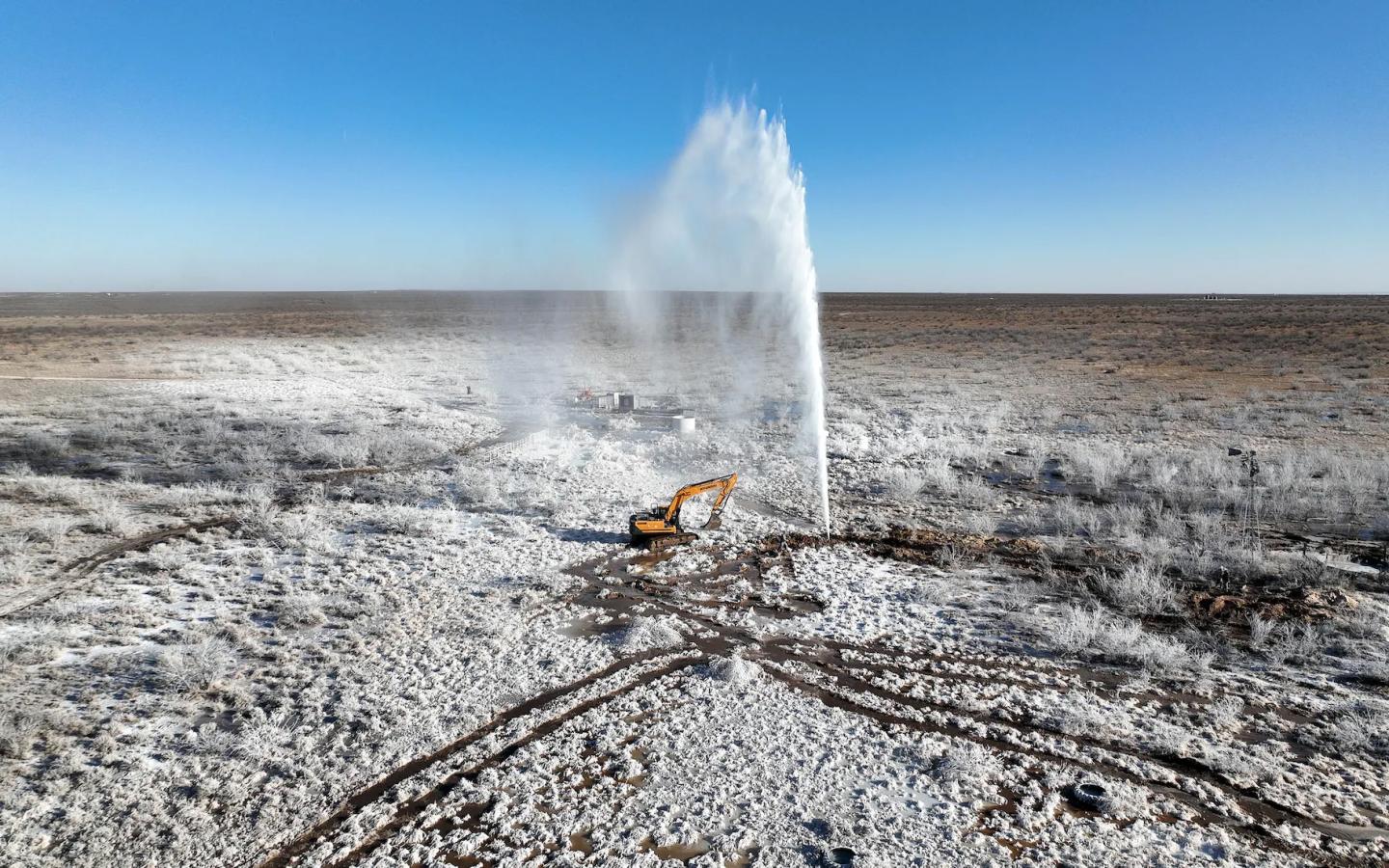 A geyser of salt water sprays out of the ground as a crane approaches on dirt that is white from the salt