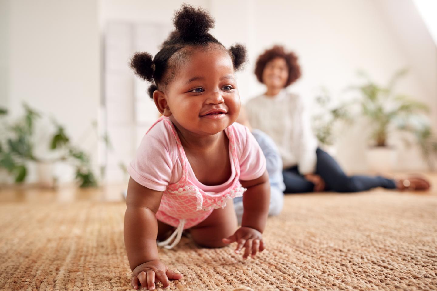 A baby crawling across the carpet with her mom watching in the background