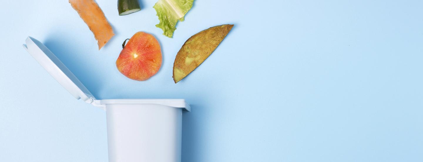 An open white trash can on a blue backdrop with food waste artfully spilled out