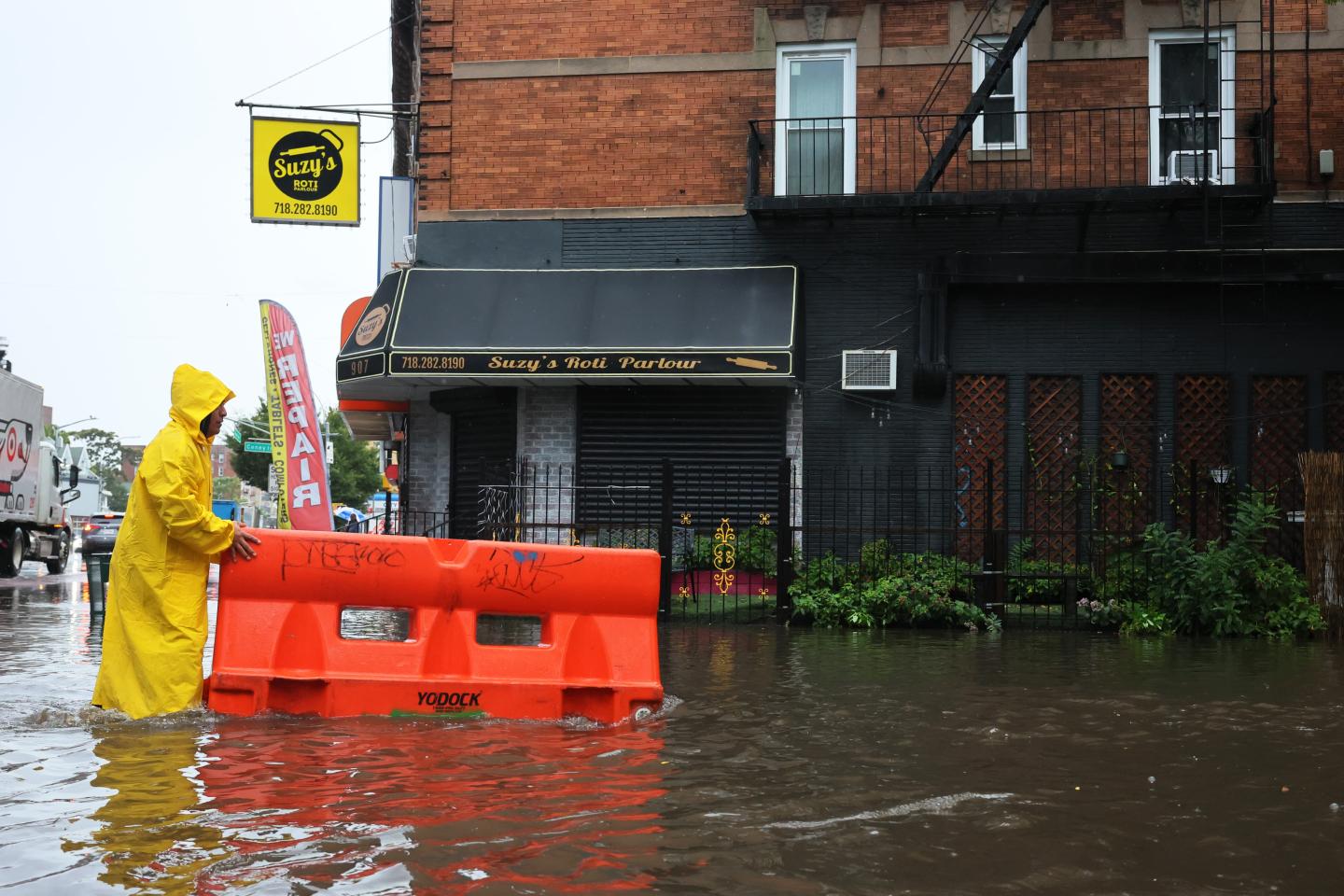 A person in knee deep flood waters wearing a yellow rain slicker moves an orange barricade