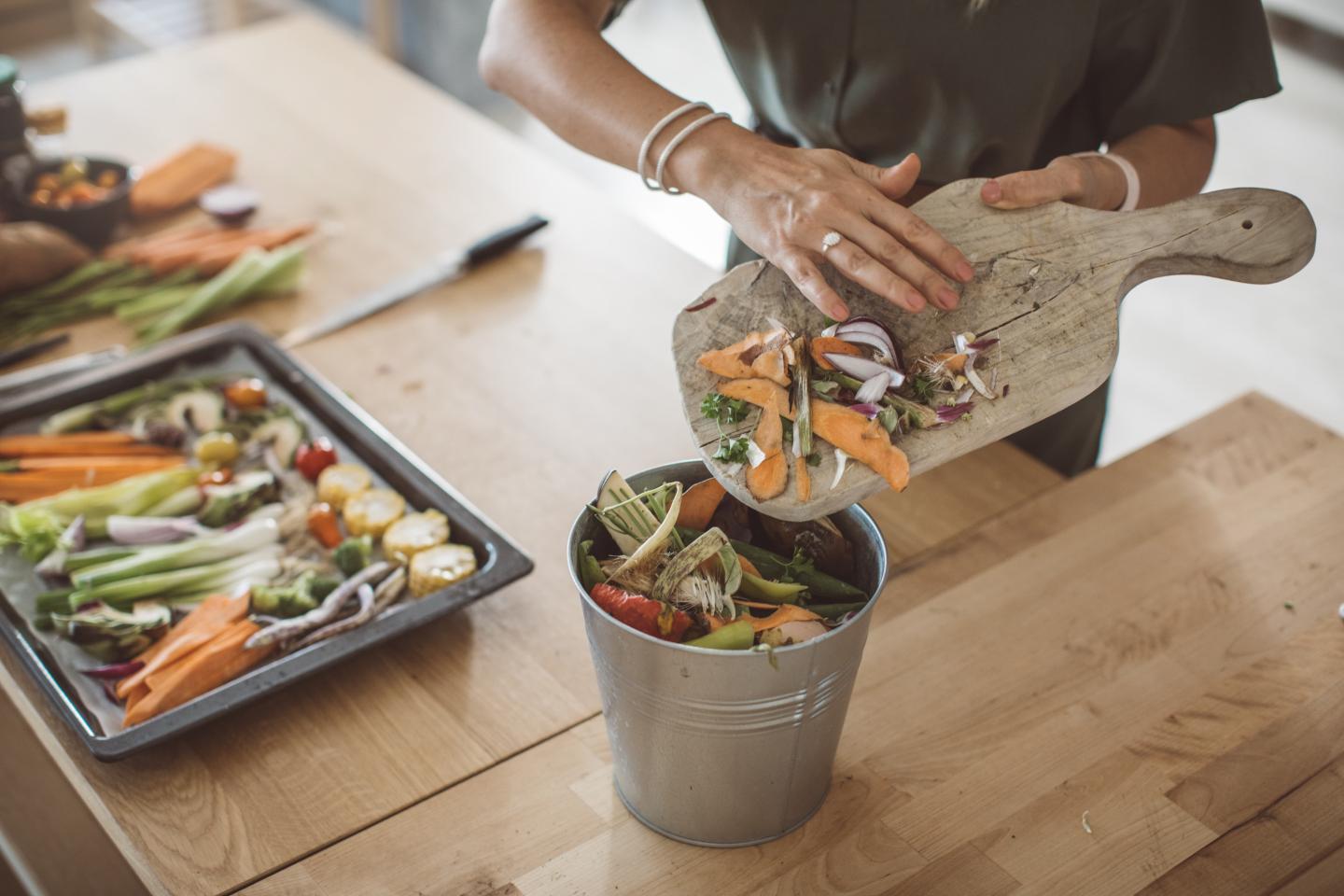 Someone pushing food scraps off a cutting board into a compost pail 