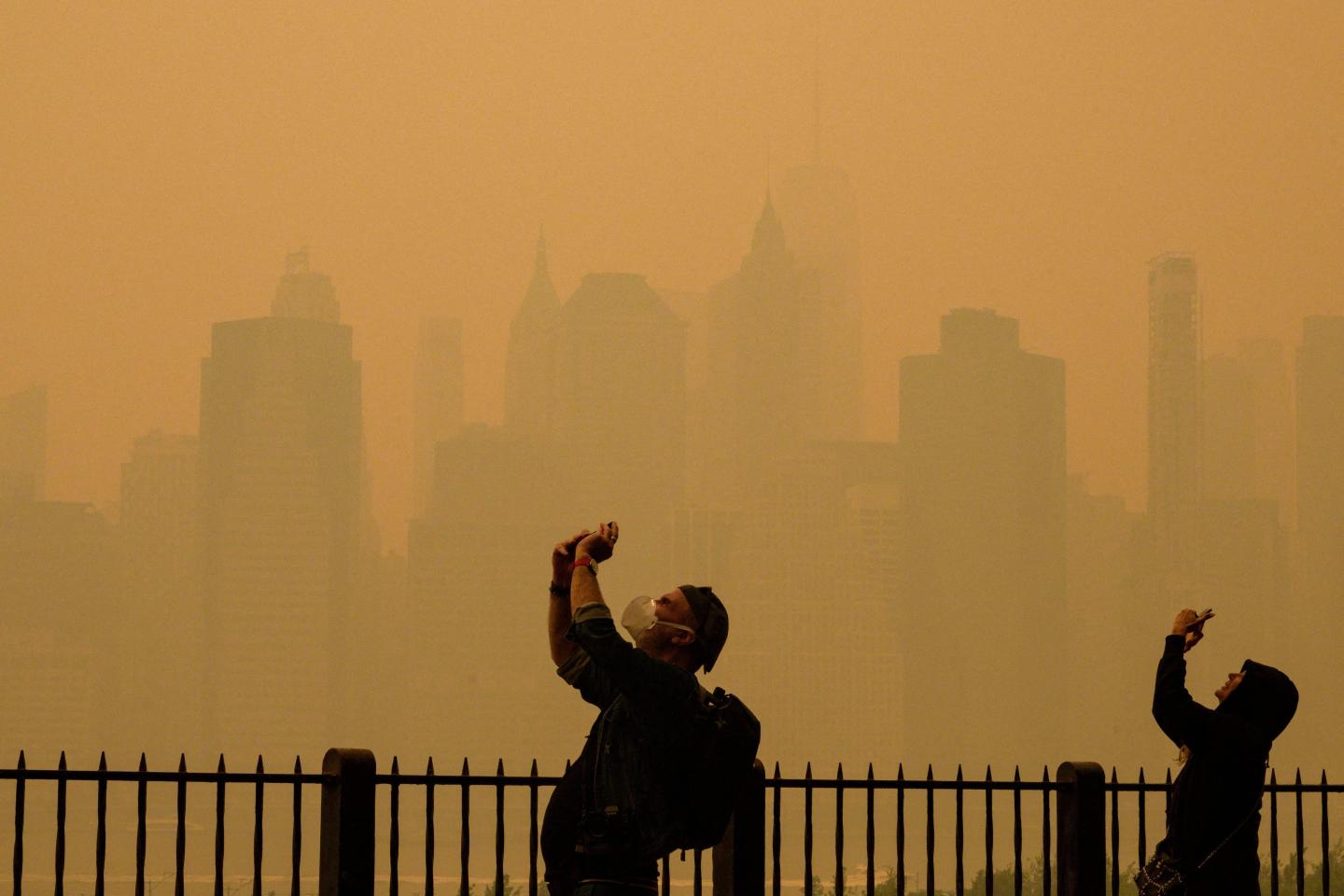 People take pictures in the foreground with an almost blocked out New York skyline in the background from deep orange haze from wildfire smoke
