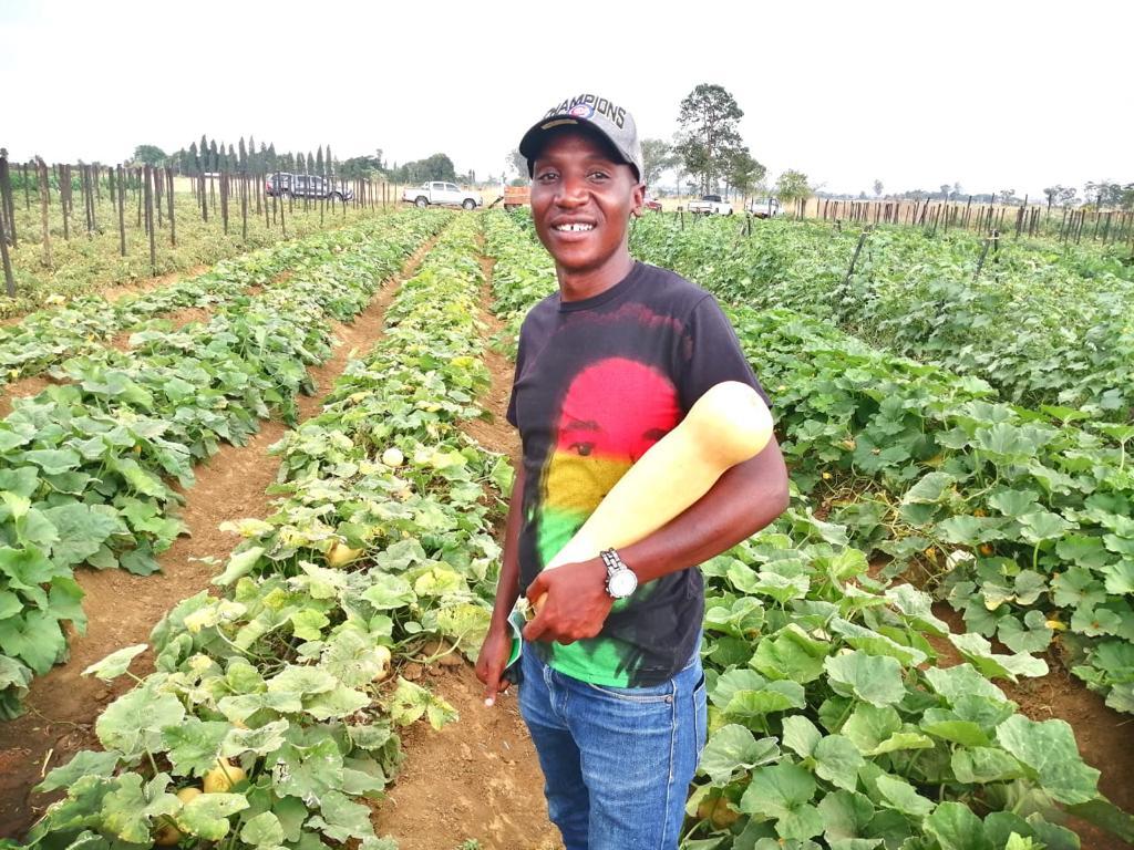 Joshua Zinzombe standing in a row of crops holding a large gourd 