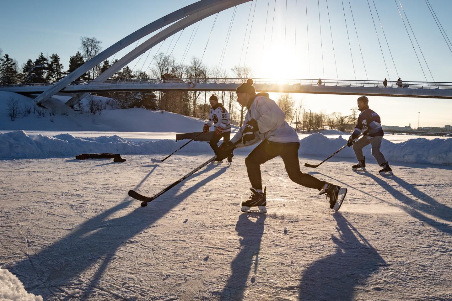 Hockey players skate across a frozen pond outdoors with a bridge in the background