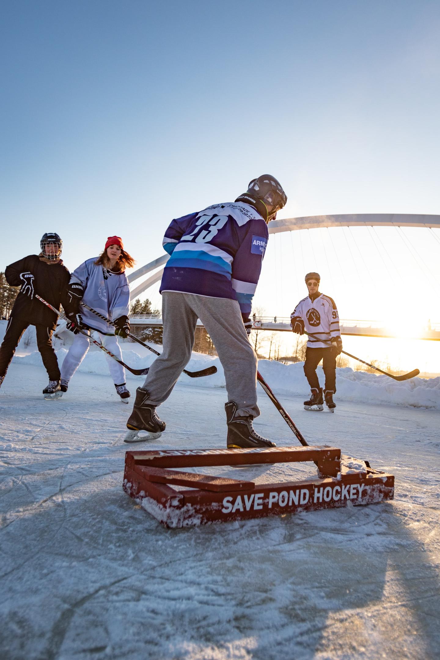 People playing hockey on an outdoor frozen pond with "save pond hockey" on a sign in the foreground