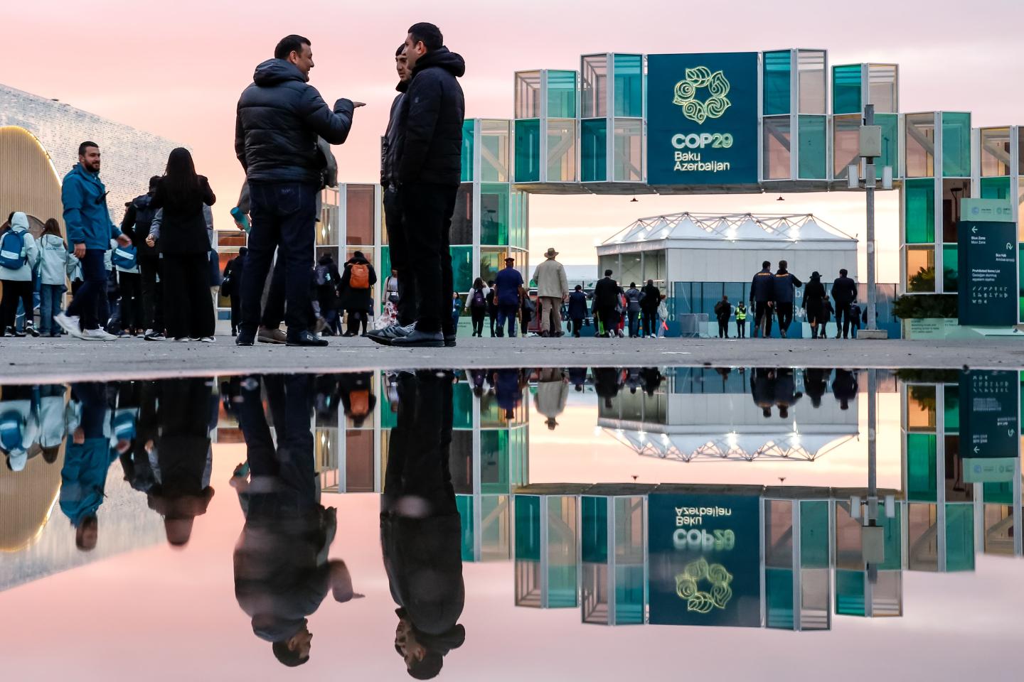 COP29 signage in the distance with a puddle reflecting both the sign and people standing nearby