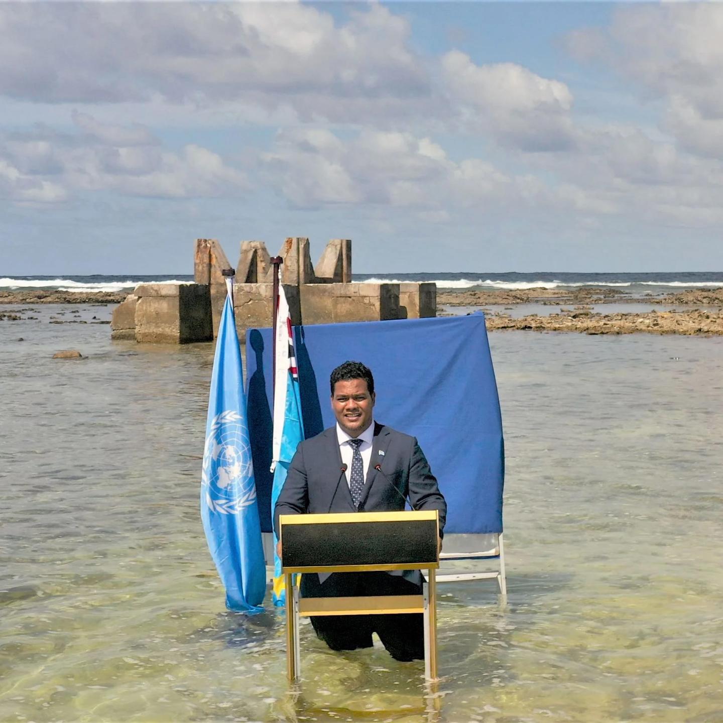 Simon Kofe holding a press conference in a suit at a lectern in knee deep water