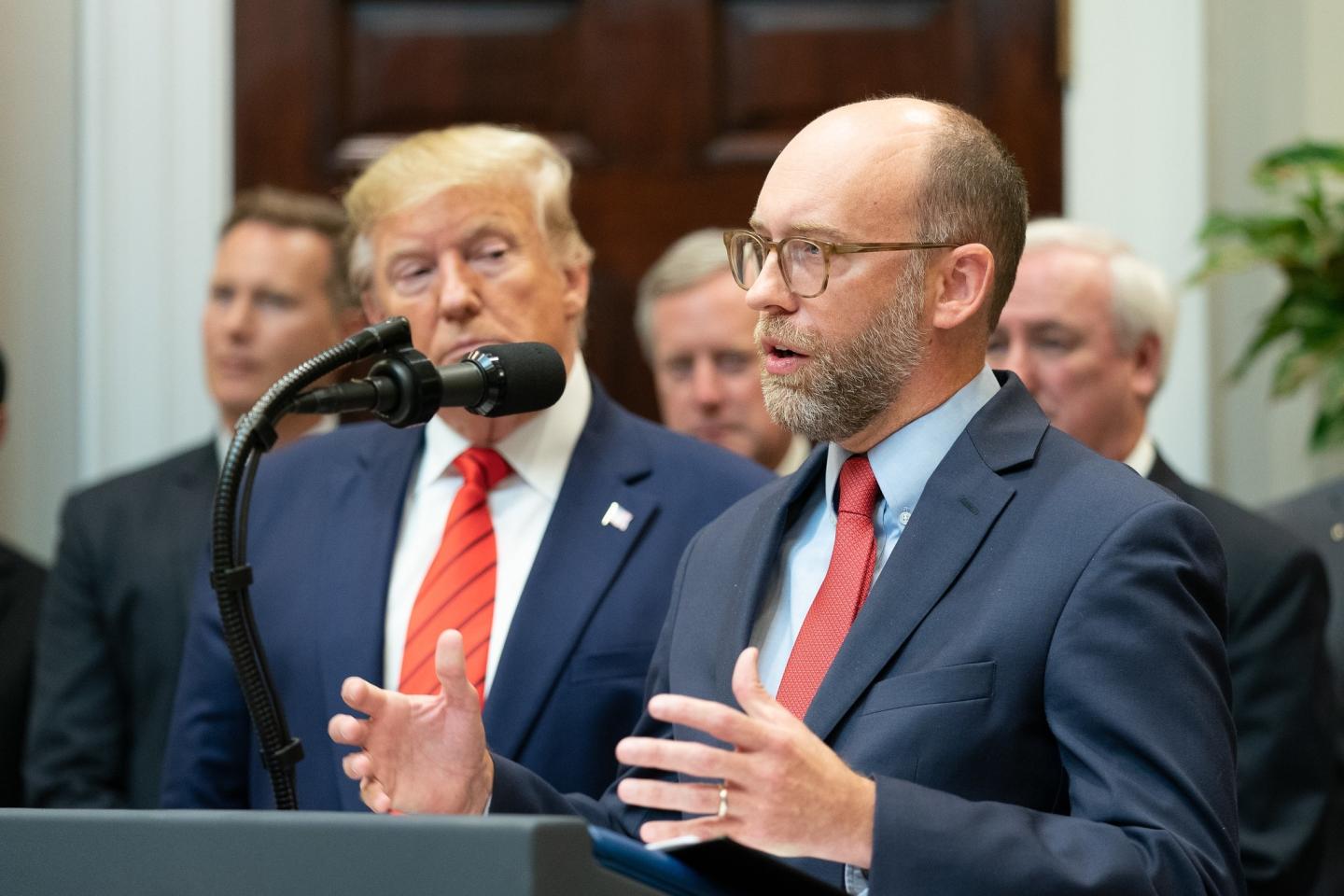 Russell Vought standing at a podium next to Donald Trump