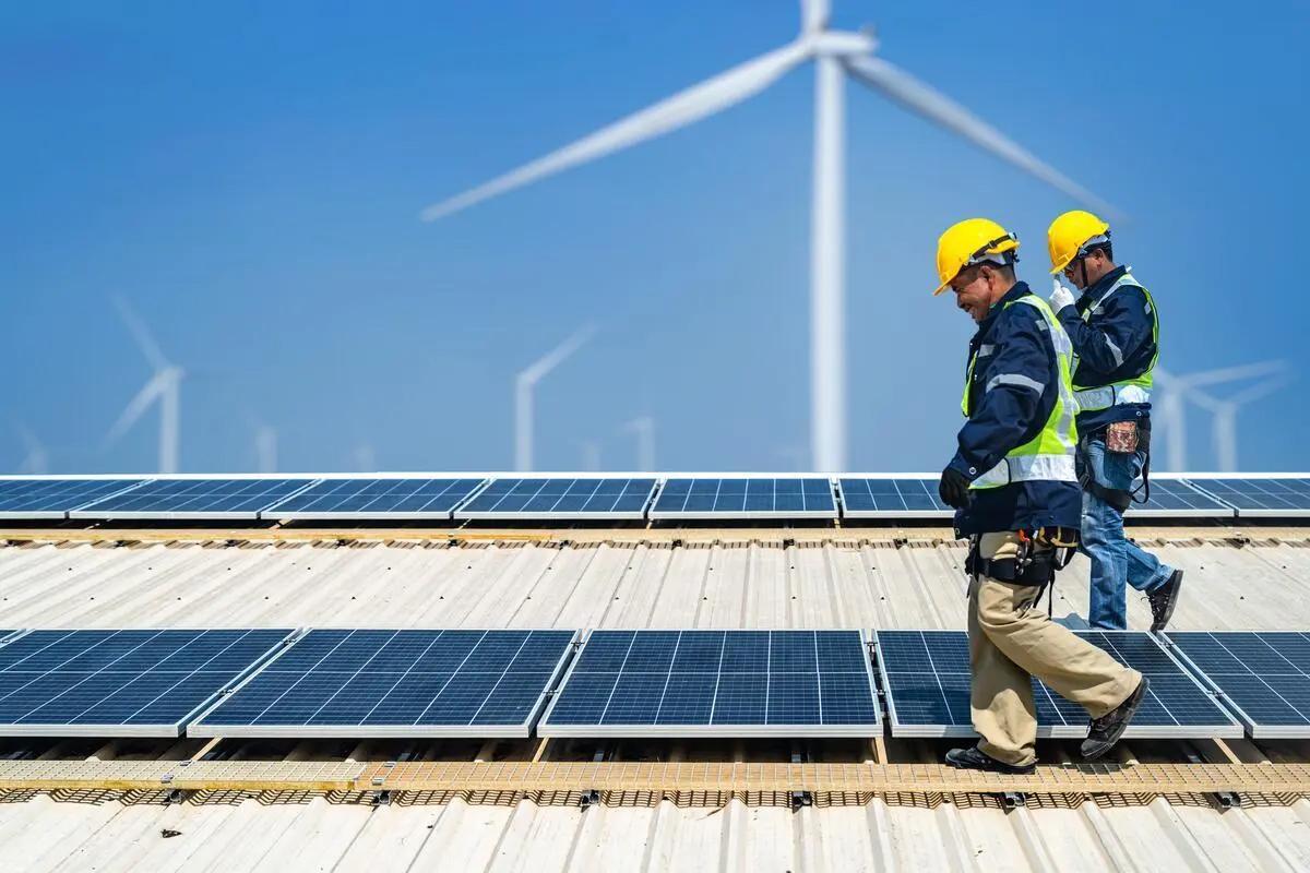 Two workers walking next to solar panels with wind turbines in the background