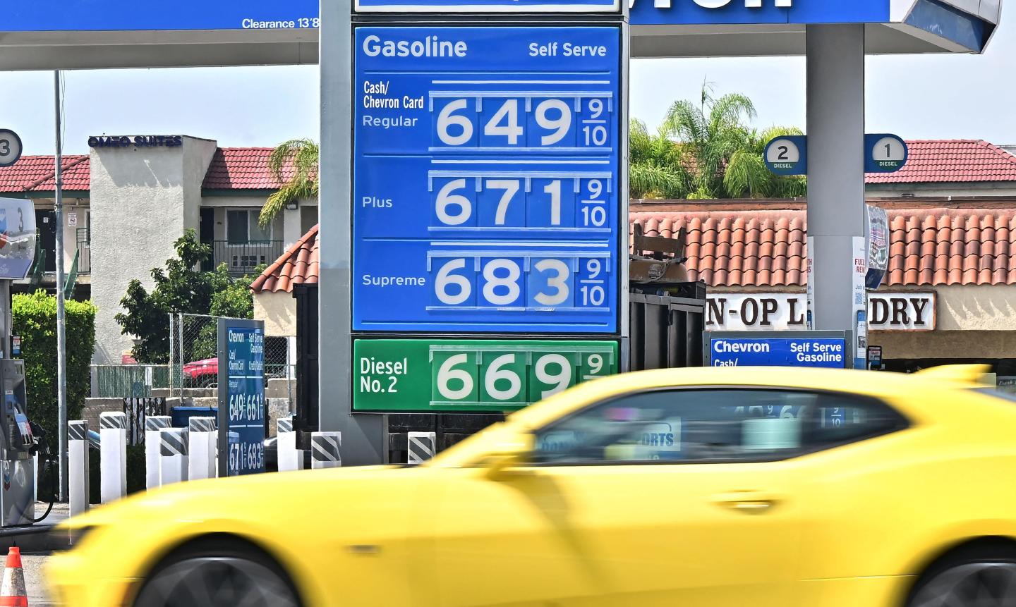 A yellow car driving in front of a gas station with gas prices starting at $6.49 per gallon
