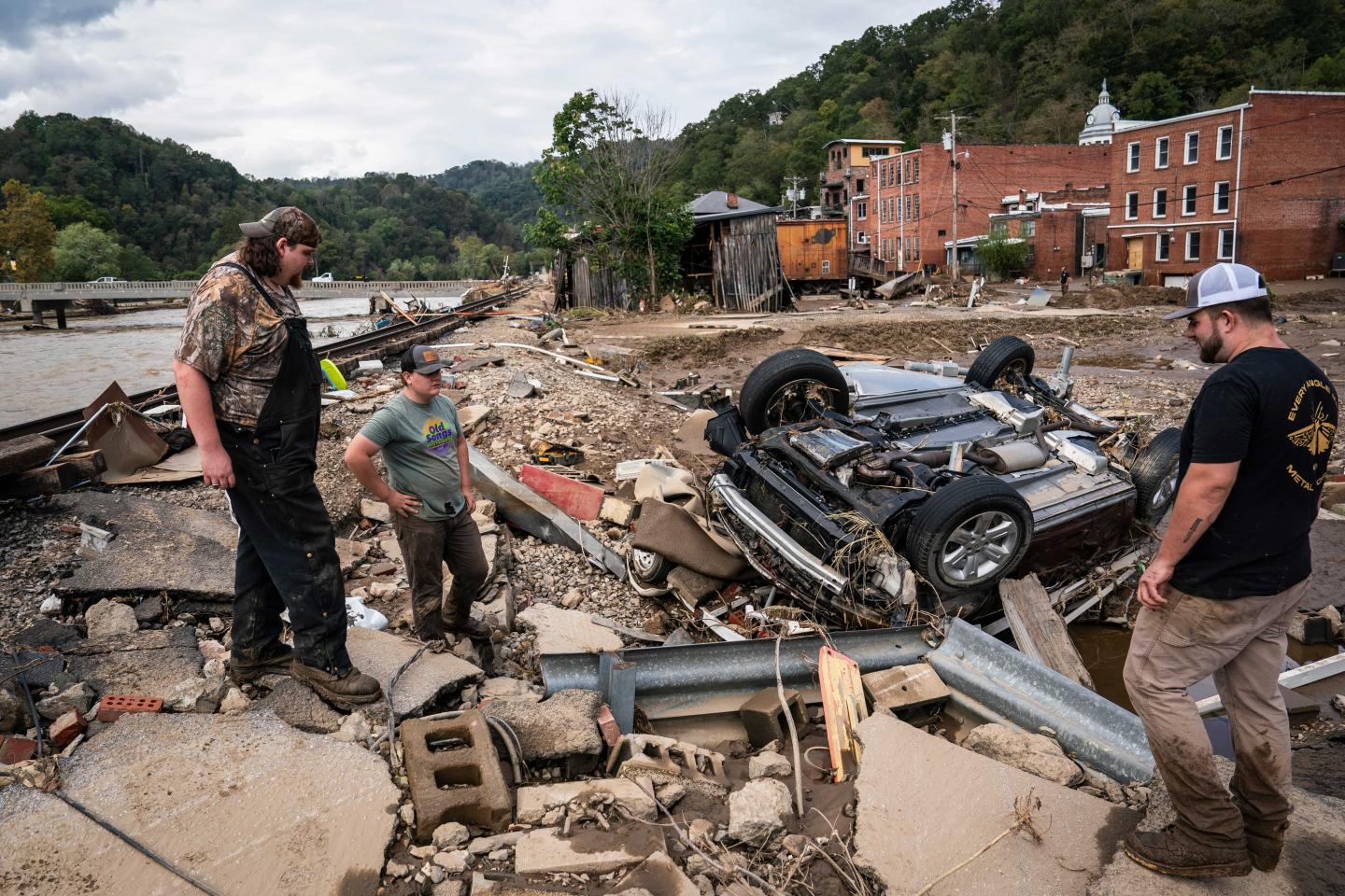 Three people standing next to an overturned and flattened car on a road that has been completely destroyed from a hurricane