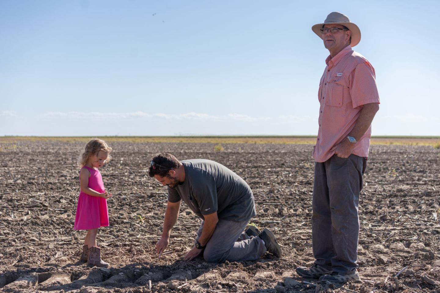 Dwane Roth standing next to his nephew Zion as he digs in the soil. His daughter is standing next to him in a pink dress