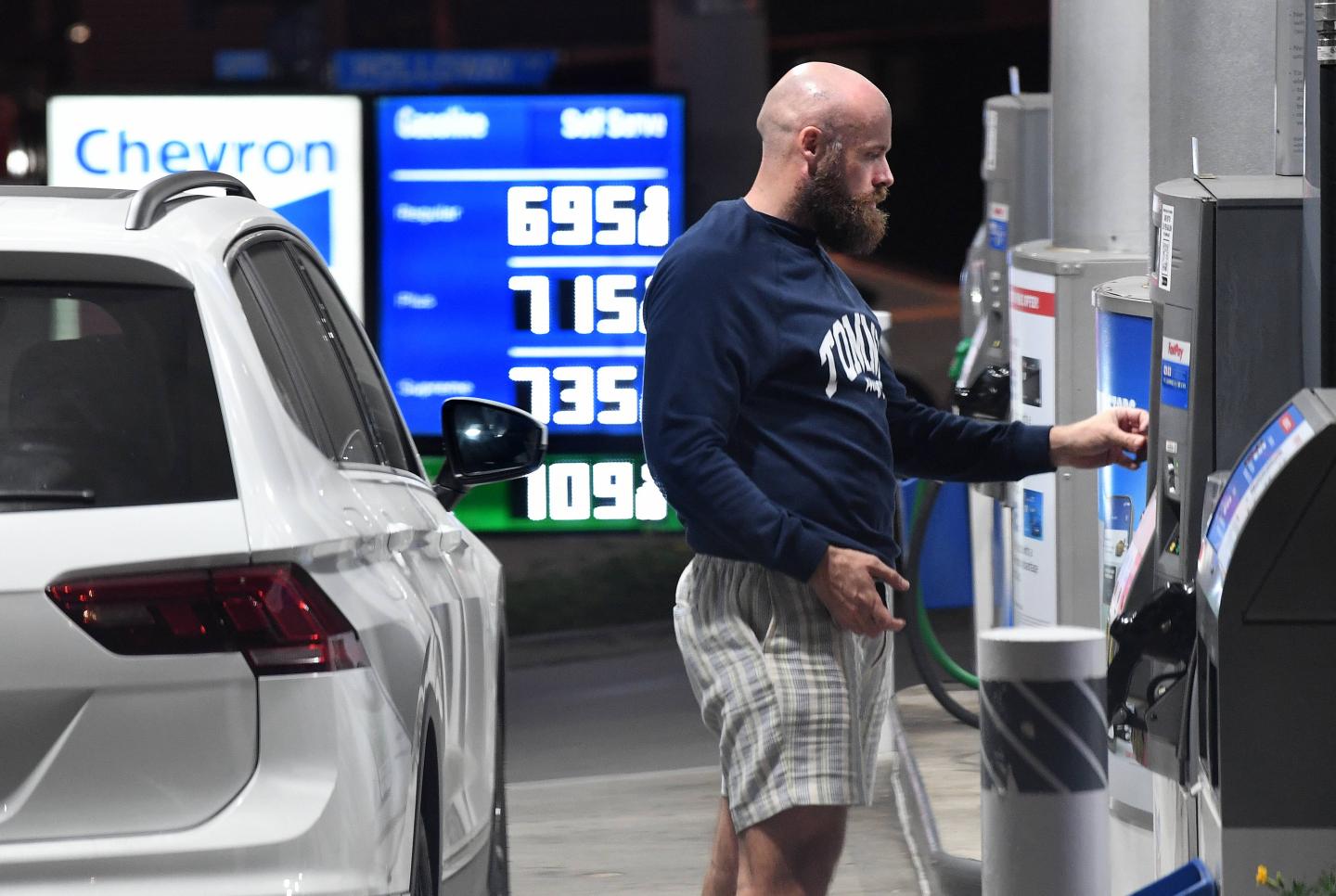 A man getting ready to pump gas, with the sign showing prices behind him starting at $6.95 a gallon