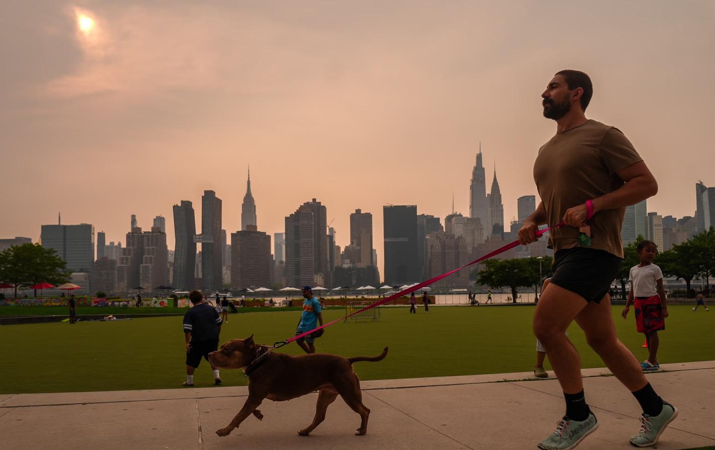 A man jogging with his dog in New York City, the skyline visible in the background, but the air is orangish and hazy from wildfire smoke