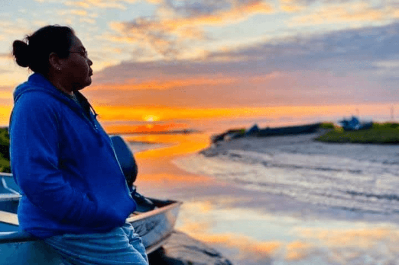 Rayna Paul sitting against a boat looking at the water while the sun sets