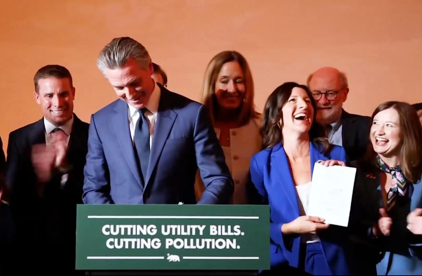 Gavin Newsom and a group of smiling people standing in front of a podium that reads "Cutting utility bills. Cutting pollution."