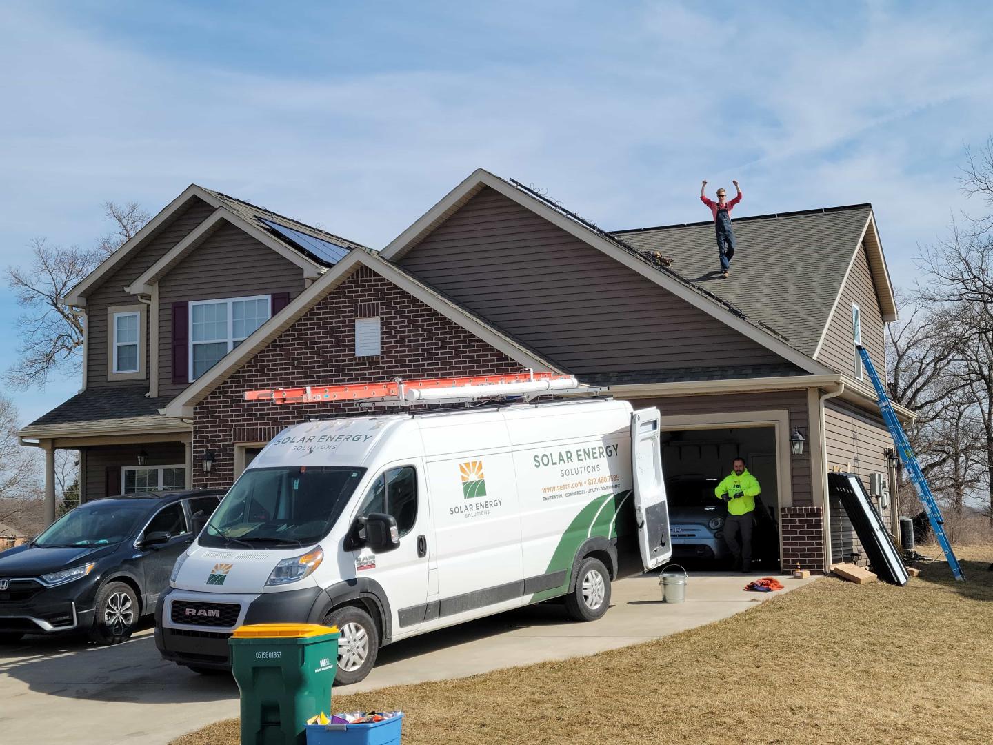 A home with solar panels being installed on it and the installer on the roof raising his arms in triumph