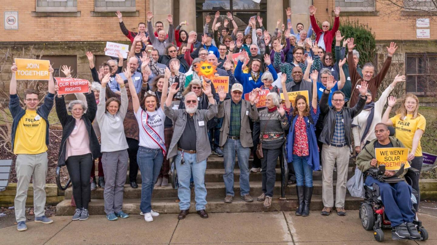 A group of people celebrating on the front steps of a building, some holding signs advocating for solar power