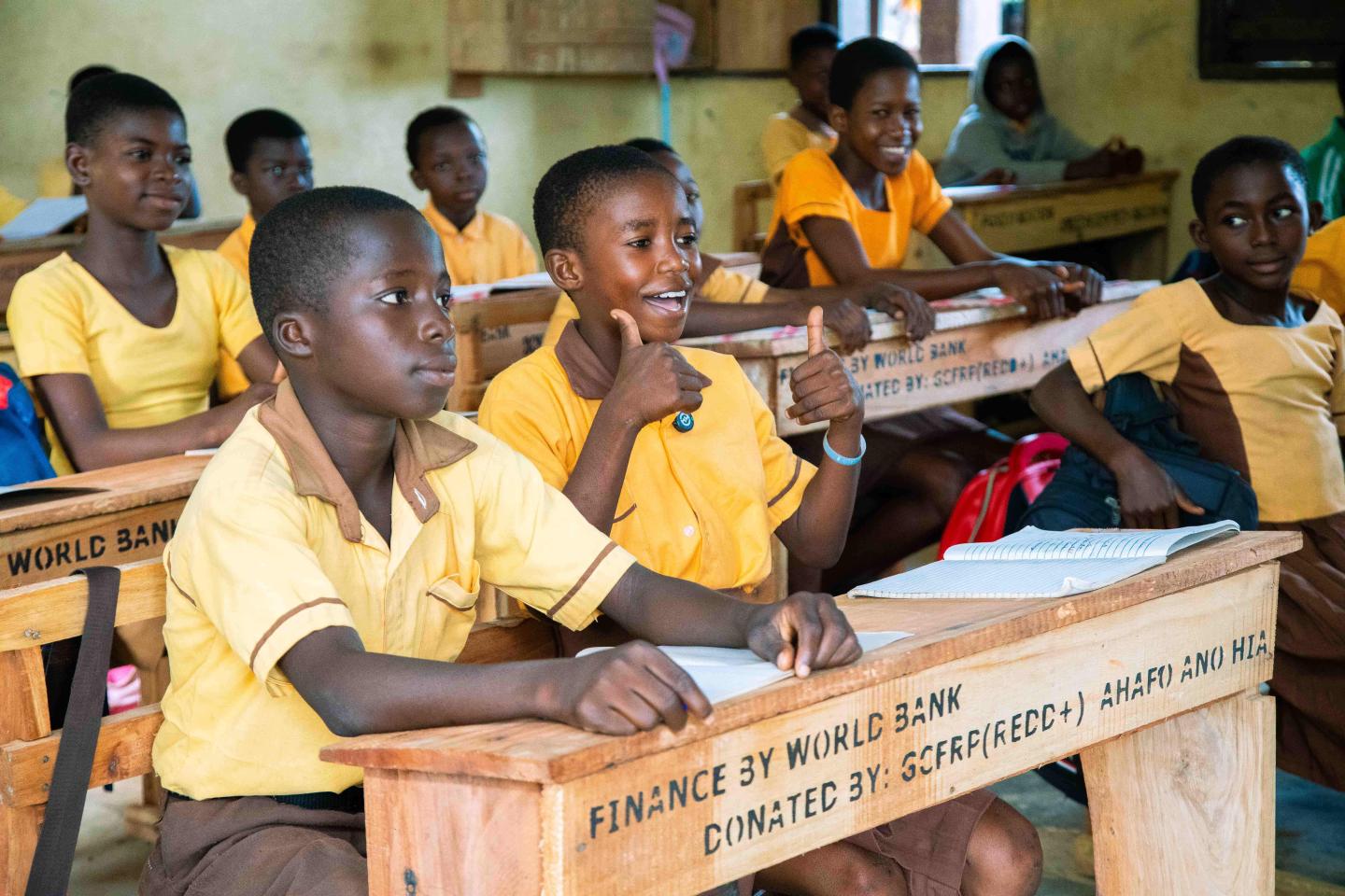 Children sitting at makeshift desks from te World Bank. One of them is smiling and giving double thumbs up