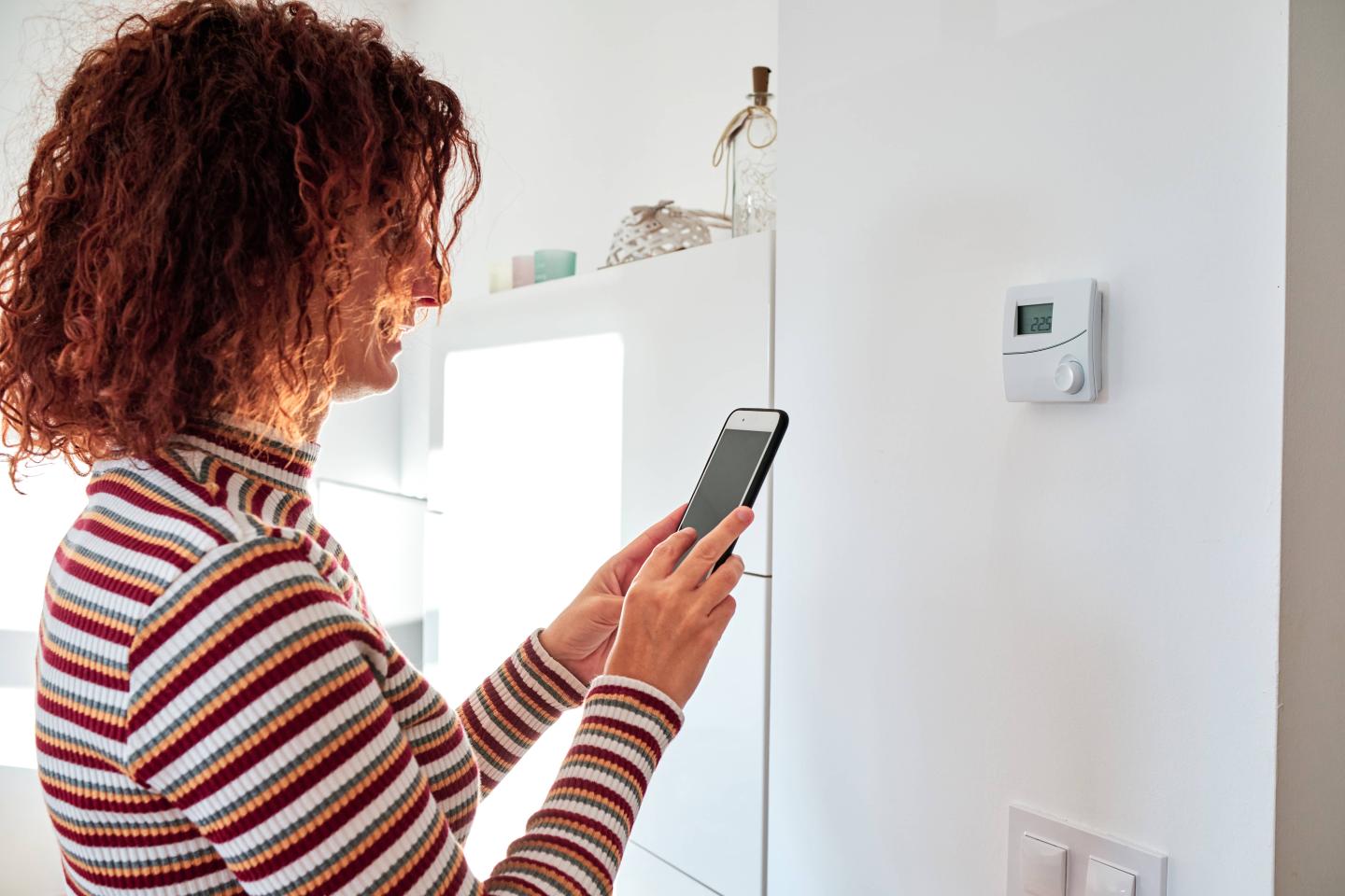 A woman checking her phone in front of her smart thermostat