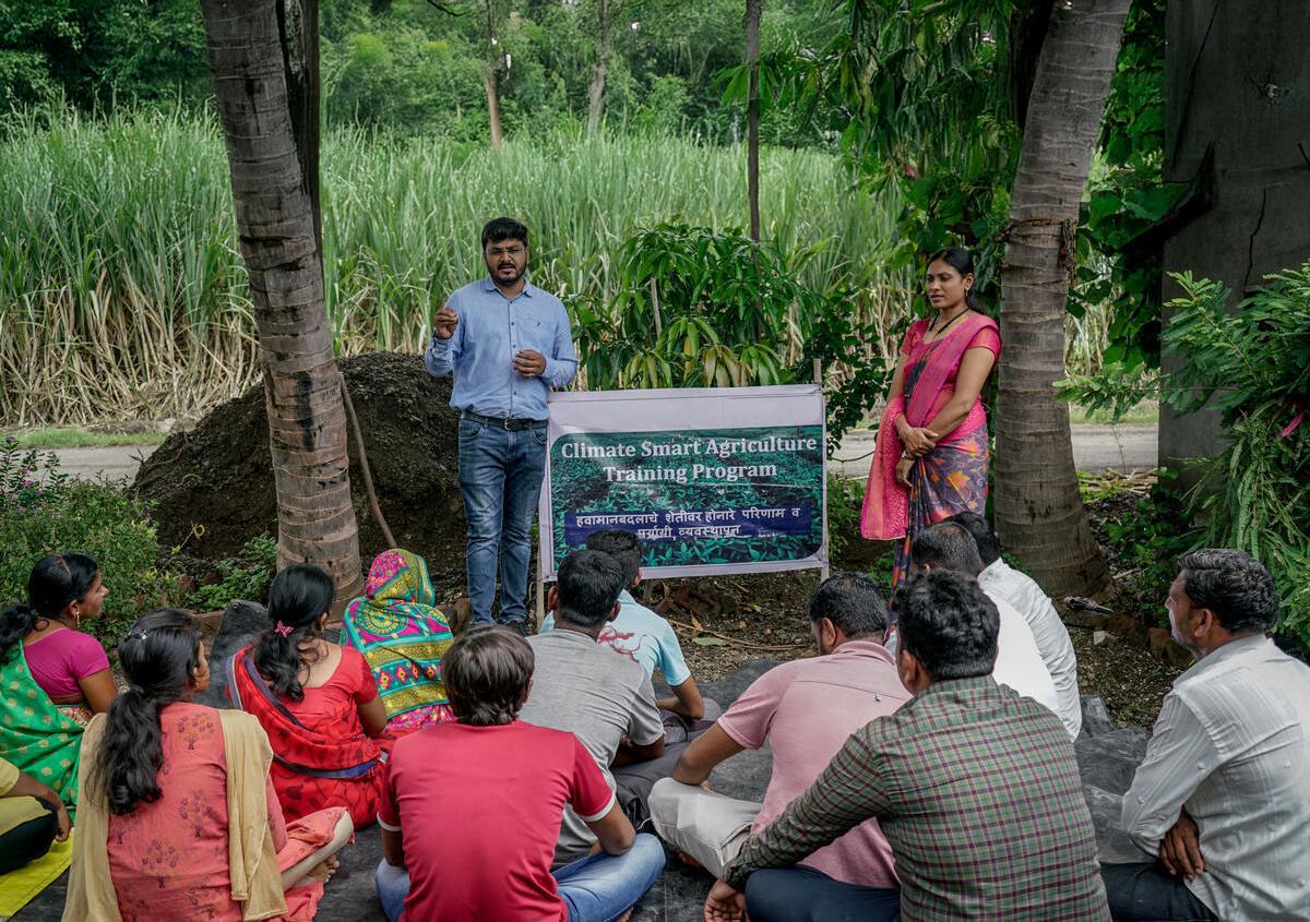Two people standing by a banner for Climate Smart Agriculture training program talking to a small crowd seated in front of them