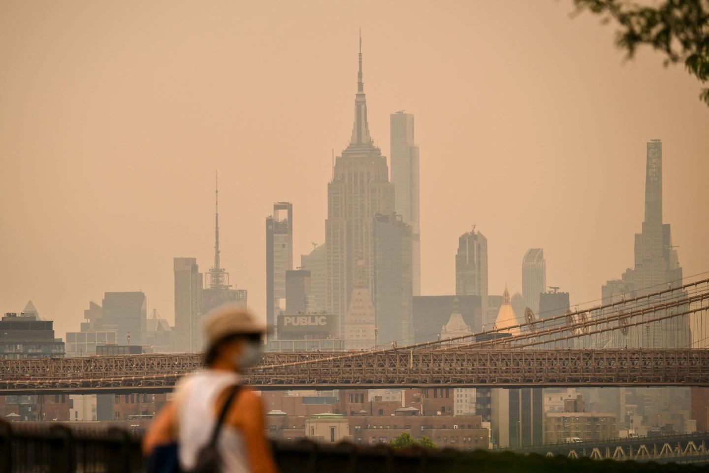 A person walking wearing a mask with the New York skyline in the background, but it is obscured in orange-ish haze from wildfire smoke