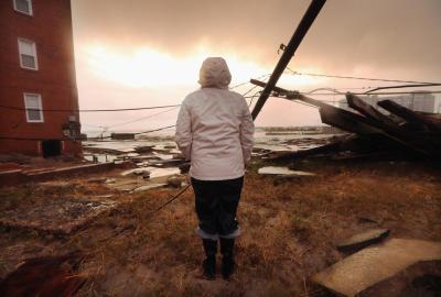 A person in a raincoat looking out over a flooded landscape with toppled telephone pole and ominous sky