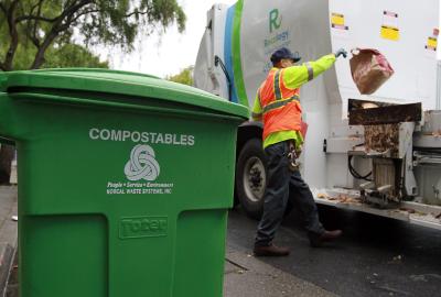 A green compostables bin sits in the foreground while a sanitation worker throws a brown paper bag into the truck 