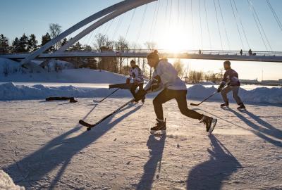 Hockey players skate across a frozen pond outdoors with a bridge in the background
