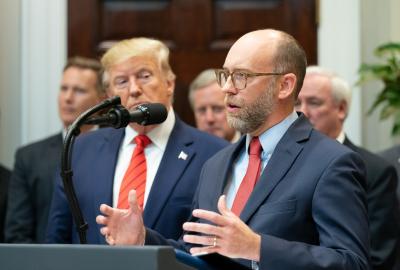 Russell Vought standing at a podium next to Donald Trump