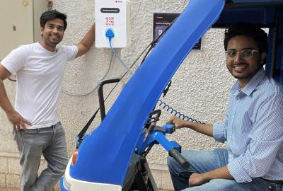 An electric rickshaw plugged in and charging with two men smiling and looking into the camera