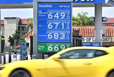 A yellow car driving in front of a gas station with gas prices starting at $6.49 per gallon
