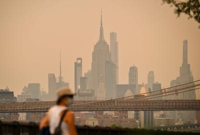 A person walking wearing a mask with the New York skyline in the background, but it is obscured in orange-ish haze from wildfire smoke
