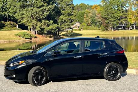a black Nissan Leaf SV plus electric car, parked on gravel beside a lake with trees in the background