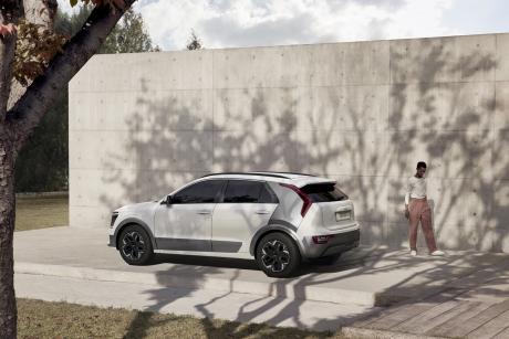 Shadows of tree branches fall over a white Kia Niro electric car, parked against a white wall. A person, dressed in white and pink, walks nearby