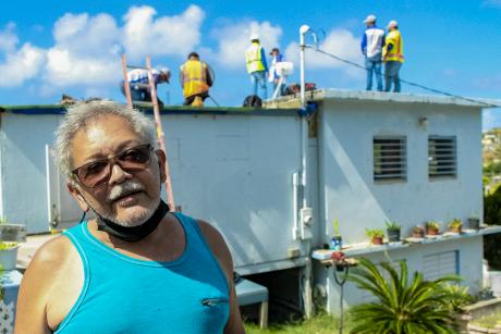 Roberto Rexach standing in front of his house while workers install solar panels on his roof