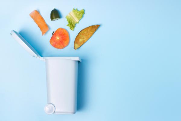 An open white trash can on a blue backdrop with food waste artfully spilled out