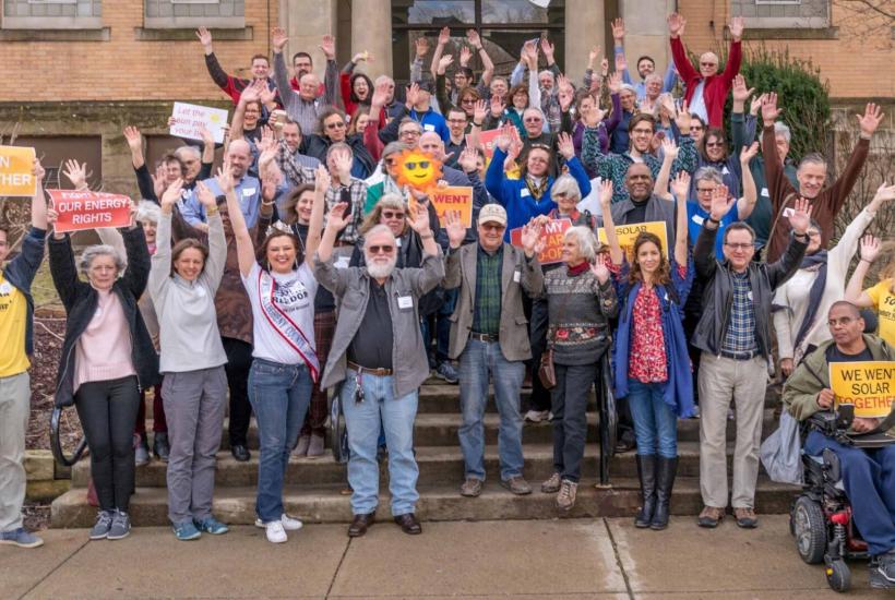 A group of people celebrating on the front steps of a building, some holding signs advocating for solar power